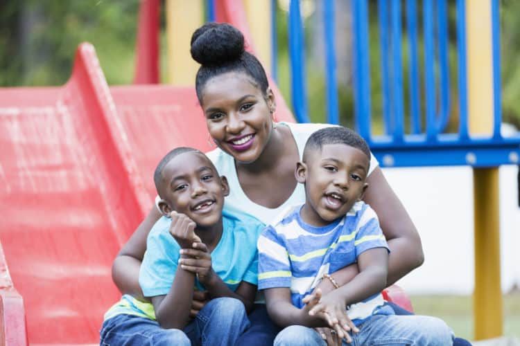 A mid adult African-American woman in her 30s sitting on a playground slide with her two son, 4 and 5 years old. The older boy on the left. They are smiling at the camera.