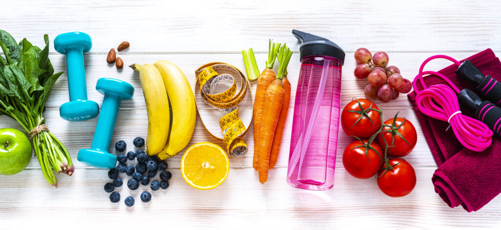 Exercising and healthy eating concept: overhead view of rainbow colored dumbbells, jump rope, water bottle, towel, tape measure and healthy fresh organic vegetables, fruits and nuts arranged side by side on white background. The composition includes spinach, tomato, carrot, banana, apple, blueberry, almonds, orange, celery, grape among others.