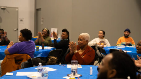 Attendees applaud and smile during the Arkansas Birthing Project Annual Meeting. Community members are seated at round tables covered with blue tablecloths in a banquet-style setting, engaged and enthusiastic as they celebrate the organization's work.
