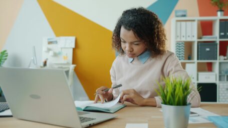 A women sitting at a desk, in front of a computer, flipping through papers with a pen in hand.