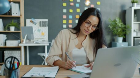 A woman sitting at a deck looks at a computer while writing something down on paper.
