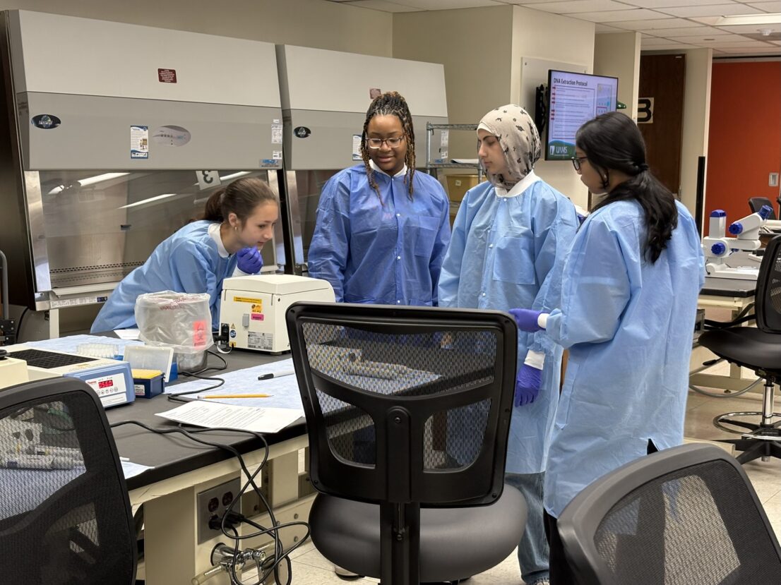 Four students in a lab setting wear protective lab coats and gloves as they look down at a small white lab device atop a table with paper, writing tools, and other lab equipment.