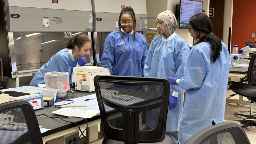 Four students in a lab setting wear protective lab coats and gloves as they look down at a small white lab device atop a table with paper, writing tools, and other lab equipment.