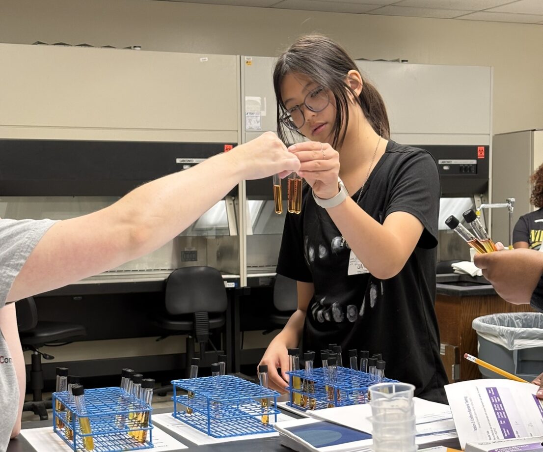 A female looks closely at two test tubes she is holding and compares it against a test tube held by someone off camera.