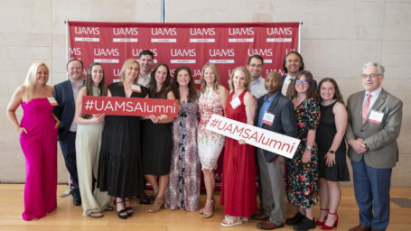 A group of people smile in front of a red UAMS Alumni background holding red and white #UAMSAlumni signs