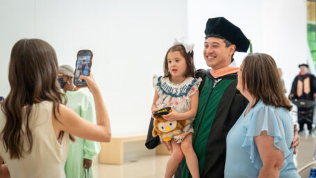 A man wearing graduation regalia poses with a toddler and woman as someone takes a picture on their phone.